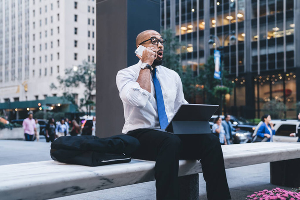 Shocked bald and bearded African American entrepreneur in white shirt with blue tie and eyeglasses sitting on stone bench and speaking on phone - Fotó, kép