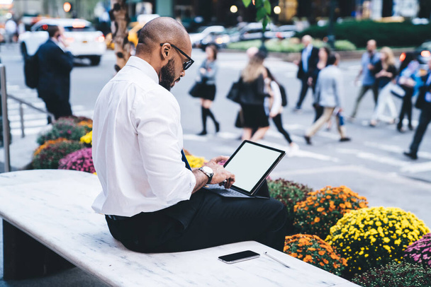 Side view of black bearded male in white shirt and black trousers using tablet with keyboard and empty screen on urban bench near flowerbed on blurred background - Fotó, kép