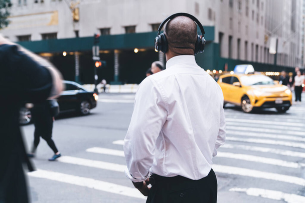 Back view of anonymous black male employee in shirt using wireless headphones listening to audio while walking against blurred crowded downtown looking away  - Фото, изображение
