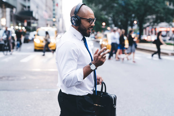 Side view of smiling elegant bald and bearded black businessman in shirt and glasses using wireless headphones enjoying music while crossing street against blurred crowded downtown  - Foto, immagini