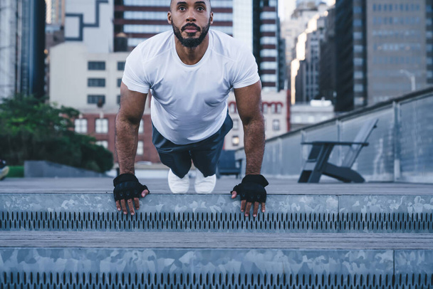 Crop from below active ethnic bodybuilder in sportswear and gloves having morning training standing in plank pose and looking away on background of buildings  - Fotografie, Obrázek