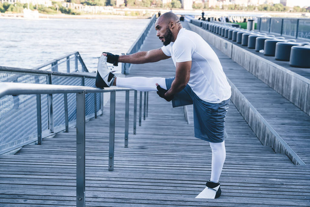Side view of bearded African American bodybuilder exercising on seafront in morning time doing legs warm up looking away on background of sea  - Foto, Bild