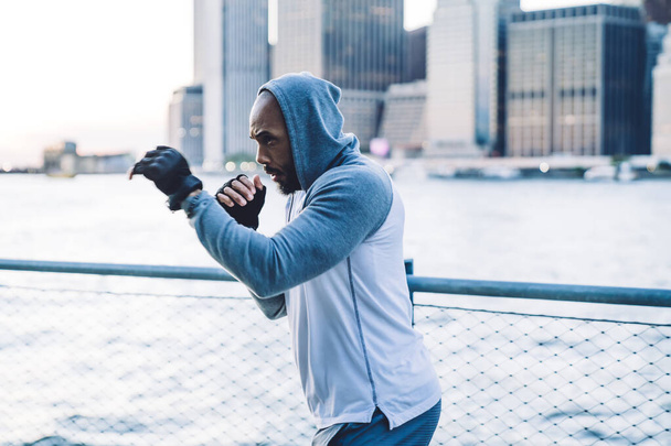 Side view of fit black man in sport costume while boxing and practicing in city on embankment on blurred background - Foto, Imagem