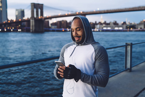 Young African American guy standing and listening to music while texting on smartphone and smiling against background of river and bridge - Foto, Bild