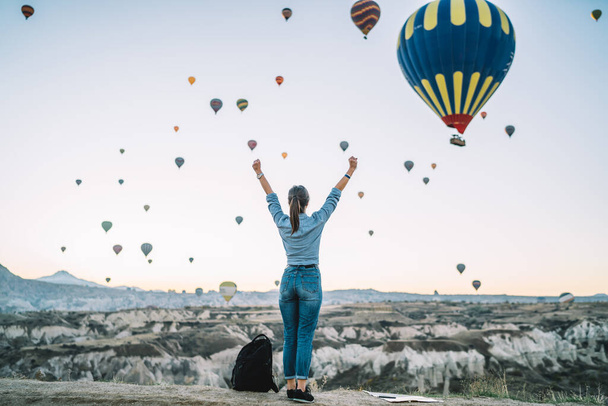 Back view of relaxed woman standing on edge of cliff in hilly terrain with hands up looking at air balloons floating in sky - Foto, immagini