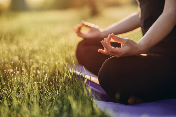 Jonge blanke vrouw beoefent yoga in lotuspositie op een vroege zonnige ochtend in een bos met gras en dauw. Gezond levensstijl concept. Ochtendmeditatie, ontspanning. Zachte selectieve focus. - Foto, afbeelding