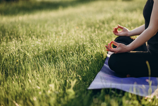 vrouw beoefent yoga in lotuspositie op een vroege zonnige ochtend in het gras met dauw. Gezond levensstijl concept. Ochtendmeditatie, ontspanning. Zachte selectieve focus. Kopieerruimte. - Foto, afbeelding