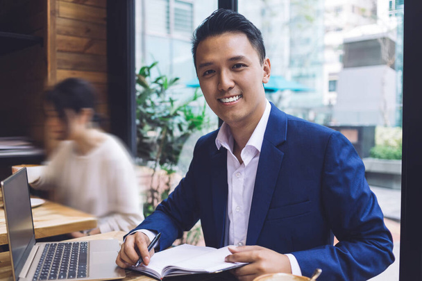 Smiling Asian man in business suit sitting at table in cafe near window with pen notebook and opened laptop and looking at camera - Фото, зображення