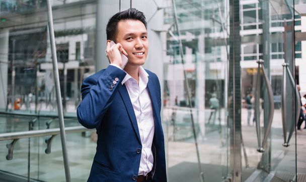 Positive Asian businessman in dark blue business suit talking on phone near entrance in glass building and looking at camera with smile - Foto, immagini