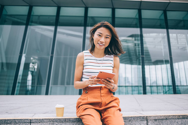 Happy attractive Asian woman looking at camera sitting on border next to coffee cup using tablet on street of Hong Kong city - Valokuva, kuva