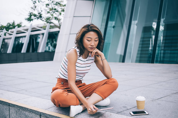Modern young Asian woman in stylish outfit watching tablet while sitting with crossed legs on street fence with coffee cup and smartphone near, Hong Kong  - 写真・画像