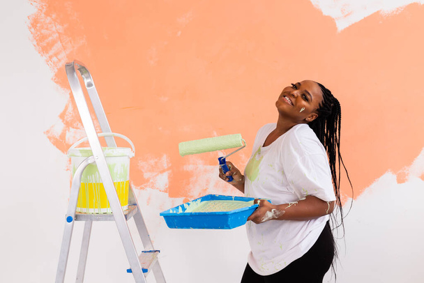 Joven mujer afroamericana feliz pintando la pared interior con rodillo de pintura en casa nueva. Una mujer con rodillo aplicando pintura en una pared. - Foto, Imagen