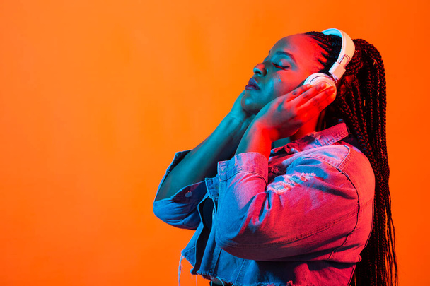 Gros plan portrait de mode beauté d'une jolie jeune femme africaine portant des dreadlocks debout sur fond orange, écoutant de la musique avec des écouteurs. Lumière au néon et espace de copie
 - Photo, image