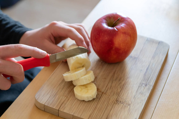 Les mains d'enfants européens coupant des fruits comme la pomme rouge et la banane avec un couteau tranchant en morceaux pour le petit déjeuner ou comme collation saine avec des vitamines sur une planche de bois sur la table de cuisine comme repas diététique
 - Photo, image