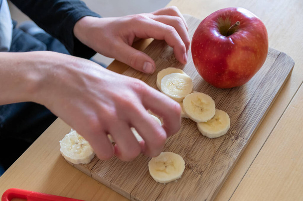 Les mains d'enfants européens coupant des fruits comme la pomme rouge et la banane avec un couteau tranchant en morceaux pour le petit déjeuner ou comme collation saine avec des vitamines sur une planche de bois sur la table de cuisine comme repas diététique
 - Photo, image