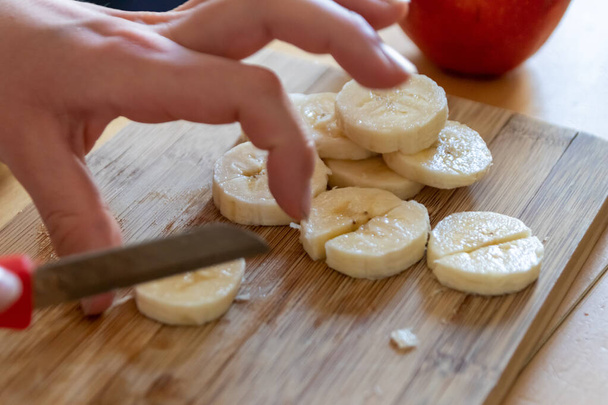 European child hands cutting fruit like red apple and banana with a sharp knife into pieces for breakfast or as healthy snack with vitamins on a wooden plank on the kitchen table as diet meal - Photo, Image