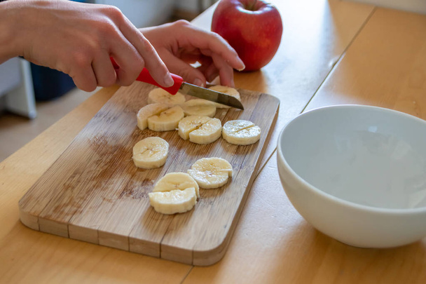 European child hands cutting fruit like red apple and banana with a sharp knife into pieces for breakfast or as healthy snack with vitamins on a wooden plank on the kitchen table as diet meal - Photo, Image