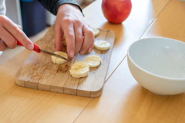Europäische Kinderhände schneiden Früchte wie rote Äpfel und Bananen mit einem scharfen Messer zum Frühstück in Stücke oder als gesunde Zwischenmahlzeit mit Vitaminen auf einem Holzbrett auf dem Küchentisch als Diätmahlzeit. - Foto, Bild