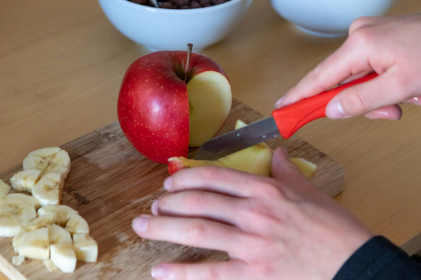 Τα χέρια του Ευρωπαίου παιδιού κόβουν φρούτα όπως κόκκινο μήλο και μπανάνα με ένα κοφτερό μαχαίρι σε κομμάτια για πρωινό ή ως υγιεινό σνακ με βιταμίνες σε μια ξύλινη σανίδα στο τραπέζι της κουζίνας ως διαιτητικό γεύμα - Φωτογραφία, εικόνα