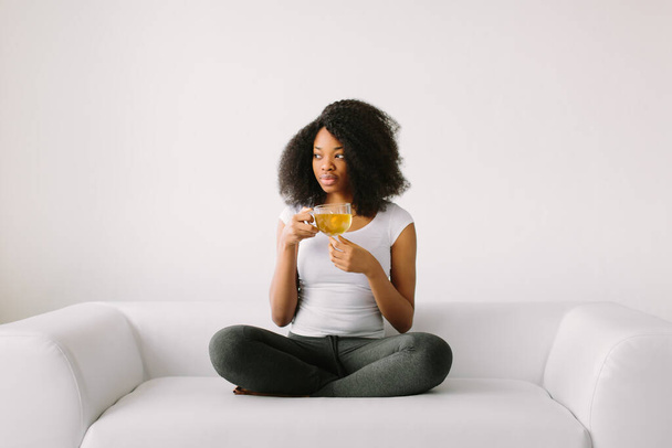 An African American young women sitting in the lotus position on white bed with cup of tea - 写真・画像