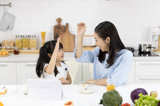 Šťastná asijská rodina Matka a Dívka jsou jíst snídani, cereálie s mlékem, Chléb s džemem a vidět digitální tablet v kuchyni doma. Zdravé jídlo a ranní aktivity - Fotografie, Obrázek