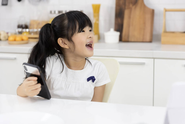Szczęśliwa Azjatka Mała dziewczynka cieszyć się smartfon podczas oczekiwania na matkę przygotowuje śniadanie w kuchni w domu. Technologia i życie codzienne - Zdjęcie, obraz