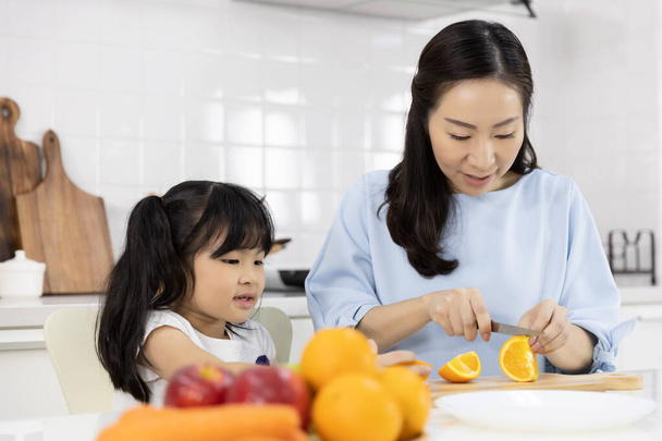 Счастливая азиатская семья Маленькая девочка наслаждается и мама готовит овощи и фрукты на кухне дома. Концепция здорового питания
 - Фото, изображение