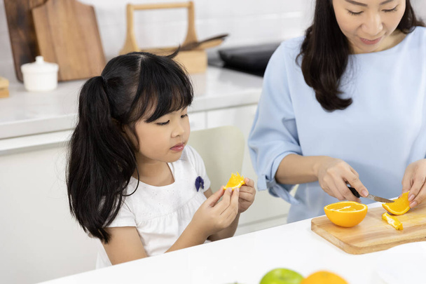 Onnellinen aasialainen perhe Pieni tyttö syö Oransseja hedelmiä ja äiti valmistaa vihanneksia ja hedelmiä keittiössä kotona. Terveellisen ruoan käsite
 - Valokuva, kuva