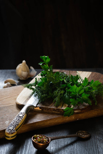 Świeże składniki do zdrowego odżywiania podczas lunchu. Pietruszka i zielenina koperkowa na drewnianej powierzchni w pobliżu noża, czosnku i pieprzu. - Zdjęcie, obraz