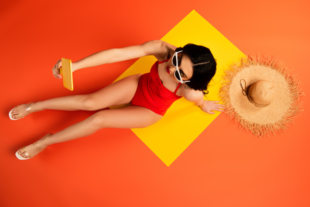 top view of young woman in sunglasses looking at mirror while sitting near straw hat on orange  - Photo, Image