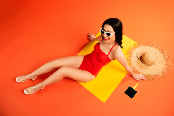 top view of cheerful woman in sunglasses and swimsuit sitting near straw hat and mirror on orange  - Fotó, kép