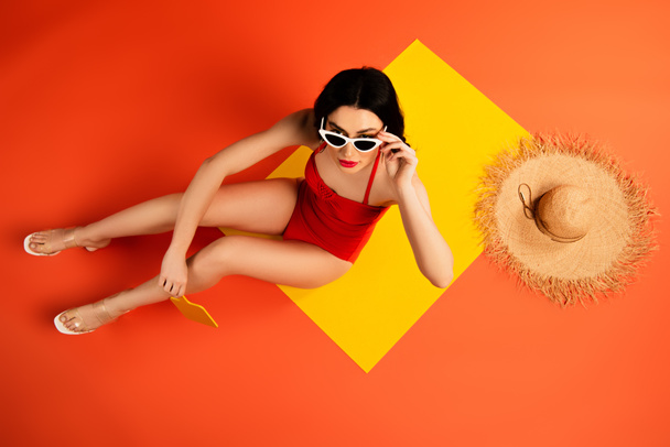 top view of woman in swimsuit touching sunglasses and sitting near straw hat and mirror on orange  - Photo, Image