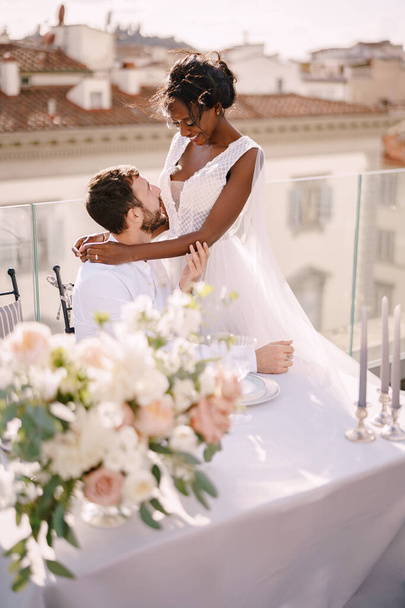 Ein gemischtes Hochzeitspaar. Traumhochzeit in Florenz, Italien. Eine afroamerikanische Braut sitzt auf dem Schoß ihres kaukasischen Bräutigams an einem Hochzeitstisch auf dem Dach. - Foto, Bild