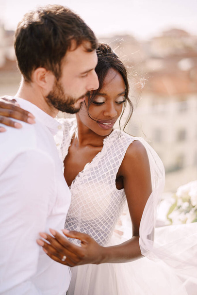 Destination fine-art wedding in Florence, Italy. Interracial wedding couple. Caucasian groom and African-American bride cuddling on a rooftop in sunset sunlight. - Zdjęcie, obraz