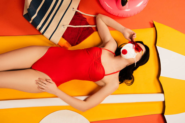 top view of beautiful woman in sunglasses, protective mask and swimsuit lying near bag and paper umbrella on orange  - Foto, immagini
