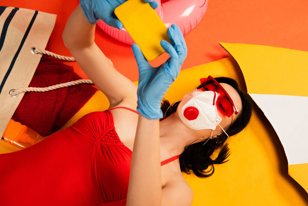 woman in sunglasses, medical mask, latex gloves and swimsuit taking selfie near bag on orange - 写真・画像