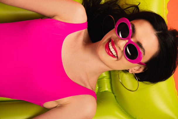 top view of cheerful girl in pink sunglasses and swimsuit lying on green inflatable mattress  - Фото, зображення