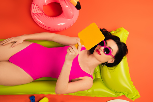 top view of woman in sunglasses and swimsuit lying on inflatable mattress and looking at mirror on orange - Fotoğraf, Görsel