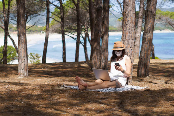 внештатная женщина, работающая в отпуске на ноутбуке, сидит в сосновом лесу на берегу моря с защитой лица от коронавируса, умной темы рабочего образа жизни и выбора жизни
 - Фото, изображение