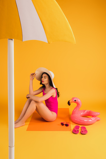 attractive woman in straw hat and swimsuit sitting near sunglasses, flip flops, inflatable ring and paper umbrella on yellow - Photo, Image