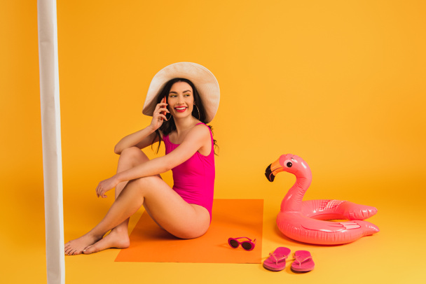 happy woman in straw hat and swimsuit sitting near sunglasses, flip flops, inflatable ring and talking on smartphone on yellow - Fotó, kép