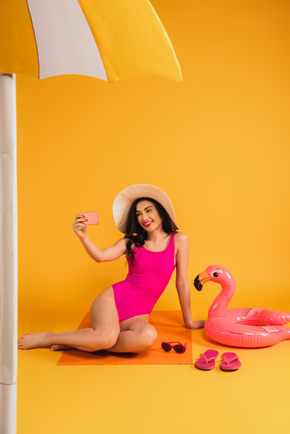 happy woman in straw hat and swimsuit sitting near sunglasses, flip flops, and inflatable ring while taking selfie on yellow - Fotoğraf, Görsel