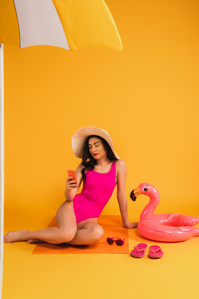 jolie femme en chapeau de paille et maillot de bain assis près des lunettes de soleil, tongs et anneau gonflable tout en utilisant smartphone sur jaune
 - Photo, image
