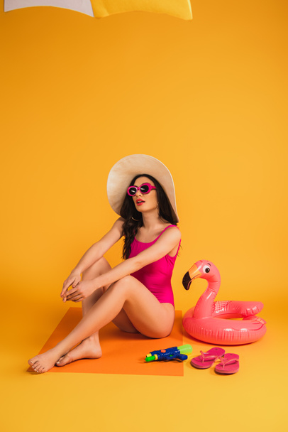 young woman in straw hat, sunglasses and swimsuit sitting near inflatable ring, flip flops and water gun on yellow - Фото, изображение