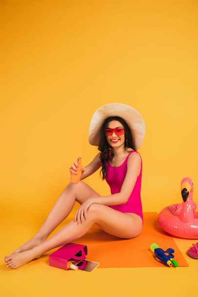 mujer feliz en sombrero de paja, gafas de sol y traje de baño sosteniendo botella con protector solar cerca de anillo inflable y pistola de agua en amarillo
 - Foto, Imagen