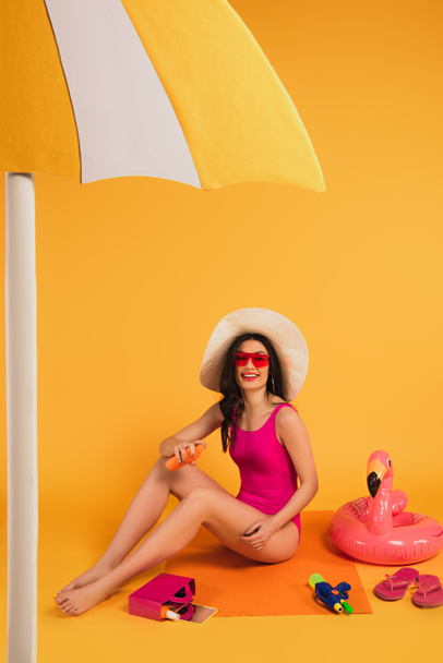 cheerful woman in straw hat, sunglasses and bathing suit applying sunscreen near inflatable ring and water gun on yellow - 写真・画像