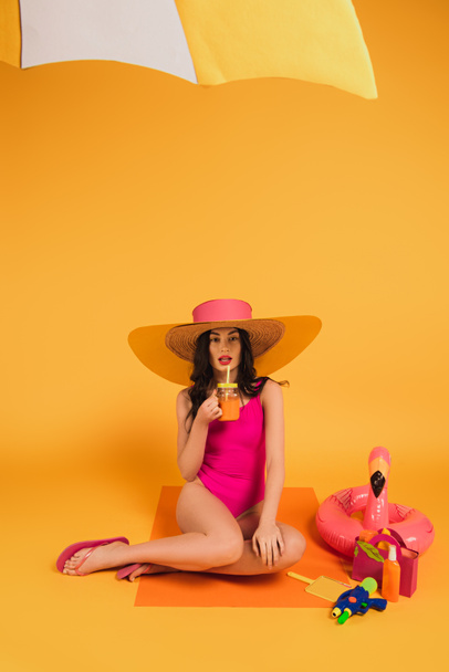 attractive girl in straw hat and swimsuit holding glass with orange juice near inflatable ring and water gun on yellow - Fotografie, Obrázek