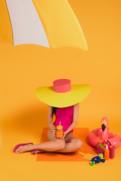 girl in straw hat and pink swimsuit holding glass with orange juice near water gun and inflatable ring on yellow - Photo, Image