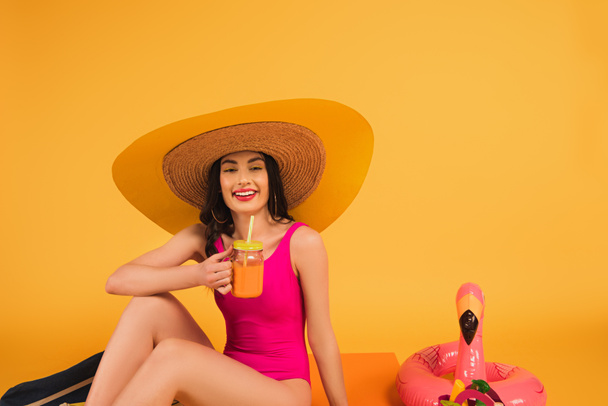 fröhliches Mädchen mit Strohhut und Badeanzug, das Glas mit Orangensaft in der Nähe von rosa Schwimmring auf gelb hält - Foto, Bild