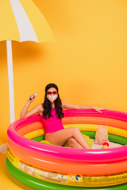 水着、サングラス、医療用マスクを着た女性が膨脹可能なプールに座り黄色の消毒剤を持っています  - 写真・画像